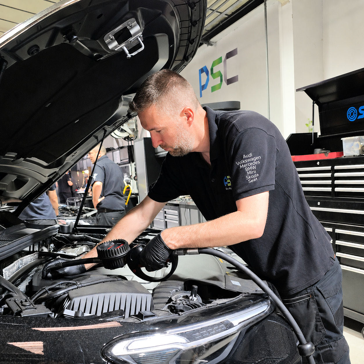 PSC Autocentre technician wearing a polo shirt listing Audi, Volkswagen, Mercedes, BMW, Mini, Skoda, Cupra and Seat, working under the bonnet of a car during a service